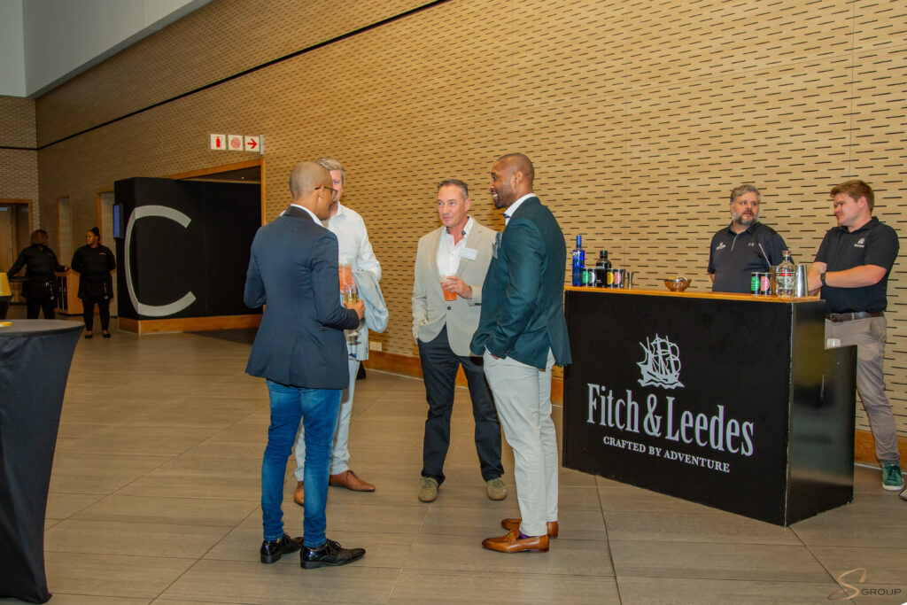 Group of casually dressed and suited men chatting with drinks at a reception near a bar labeled Fitch &amp; Leedes.