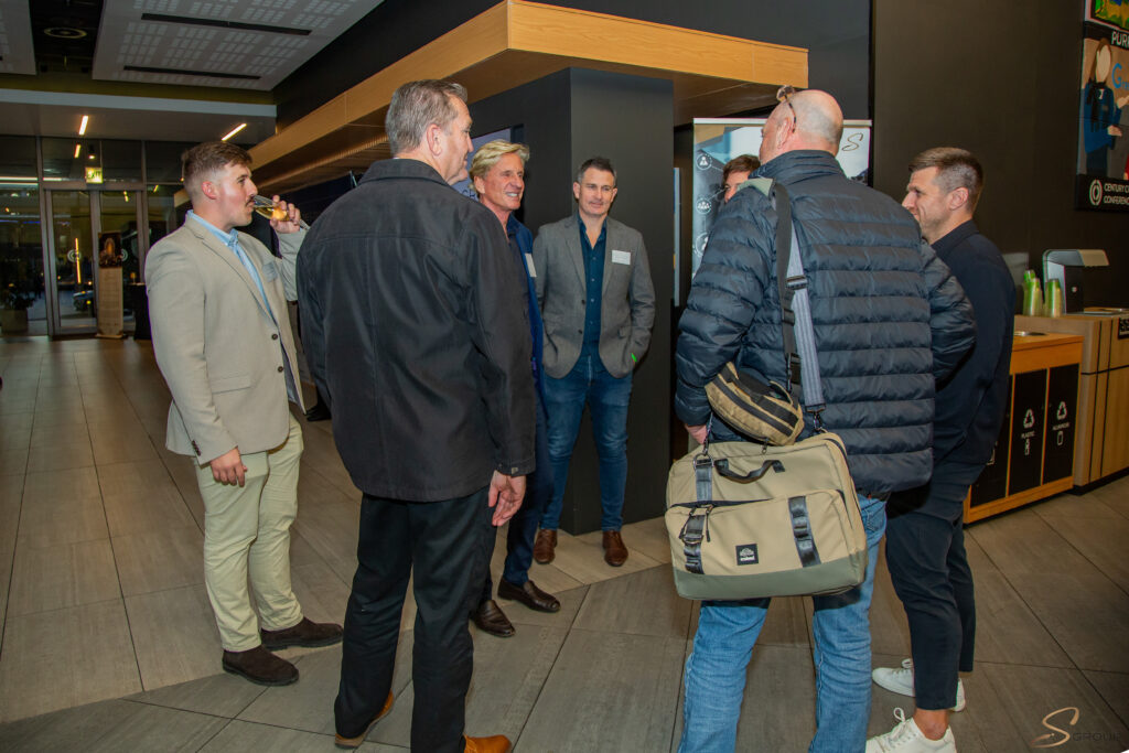 Group of men in business casual chatting in a modern conference lobby; one man drinks from a glass, a large bag in the foreground.