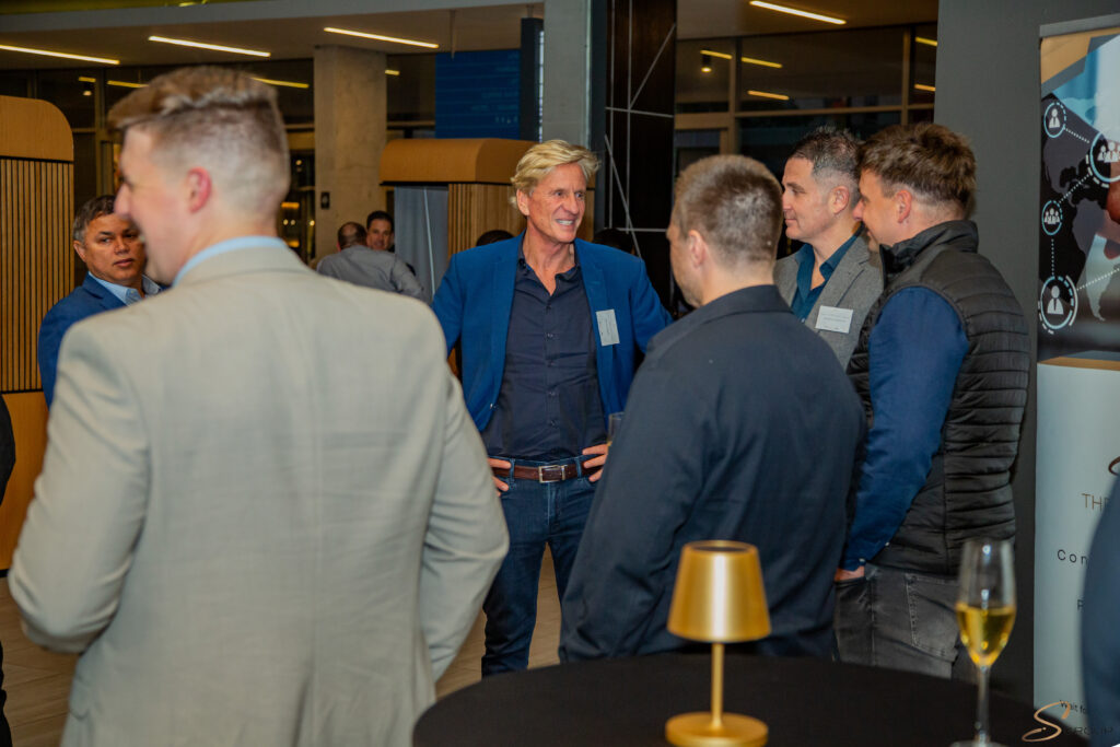 Group of professionally dressed men conversing at a networking event, name badges visible, in a modern venue with tall windows and display panels.