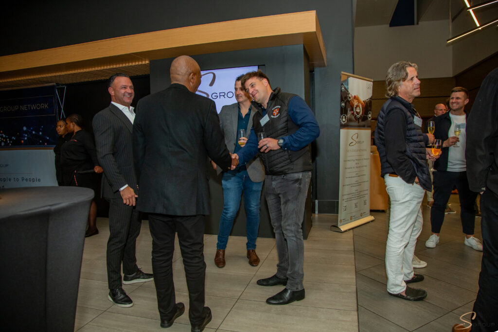 Professionals at a networking event exchanging a handshake while others chat, with banners and drinks in the background.