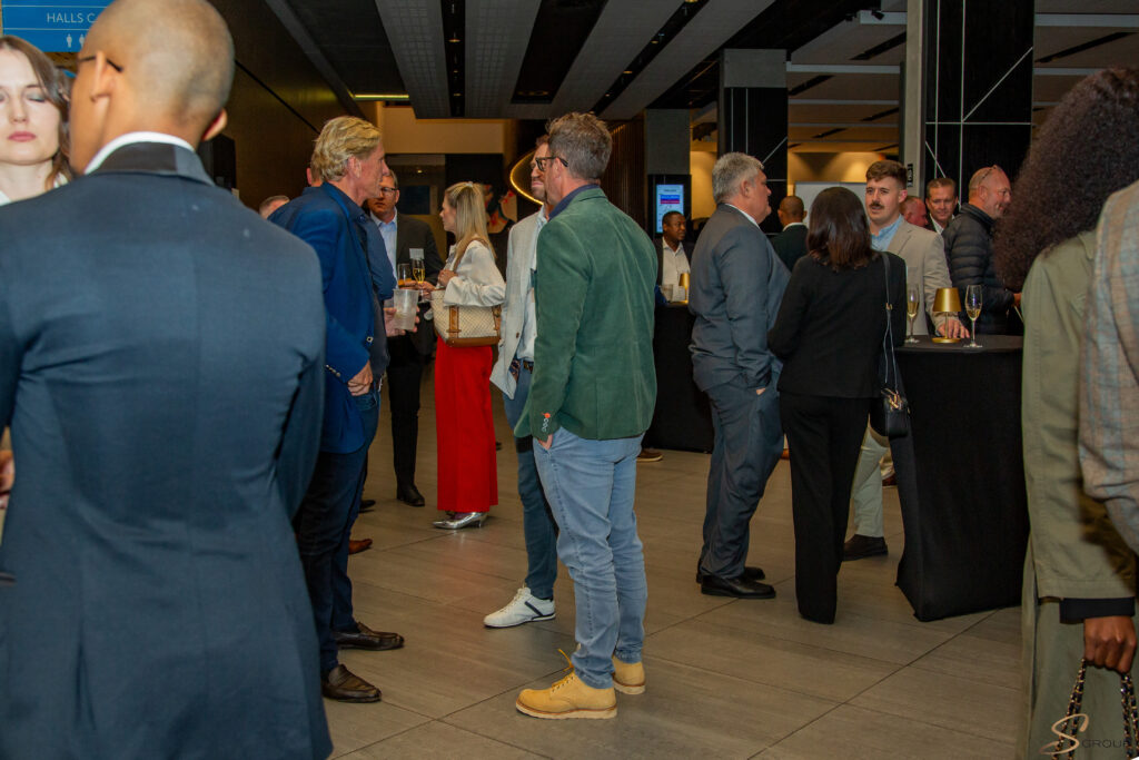 Professionals networking at a evening reception: people chat in small groups, some holding drinks, in a modern venue with dark columns and warm lighting.