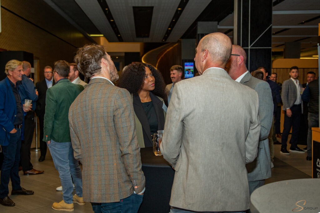 Group of professionals in smart jackets conversing at a networking event in a modern venue, some holding drinks.