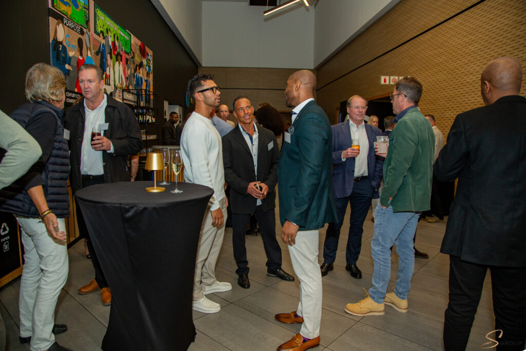 Group of professionals conversing at a standing reception, drinks in hand, around high-top tables in a modern venue with a colorful mural backdrop.