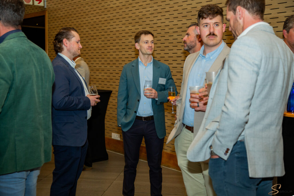 Group of men in business attire converse at a reception, holding drinks, against a wood-pattern wall backdrop.