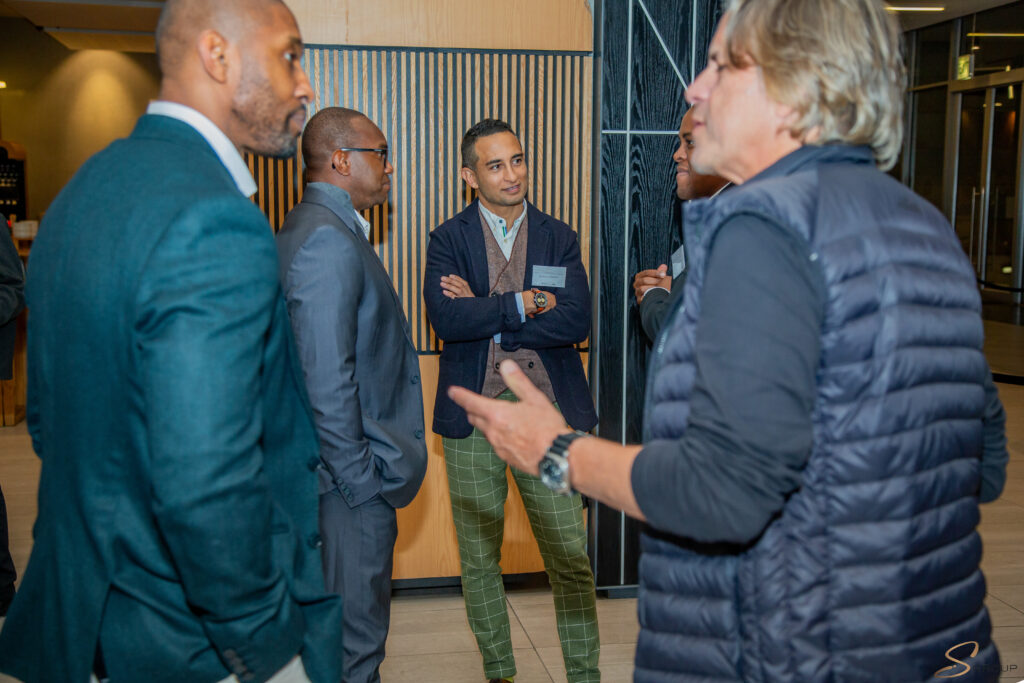 Five men in business casual wear stand in a modern lobby, engaged in conversation, with a wooden slat wall behind them.