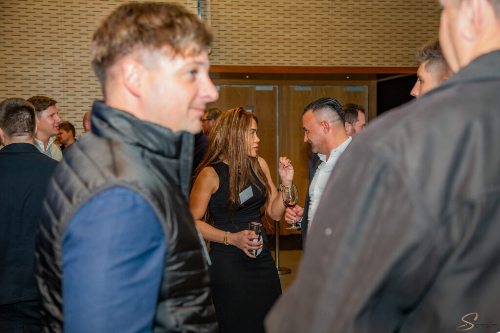 Woman in a black dress holding a wine glass talks to a man in a white shirt at a crowded social event.