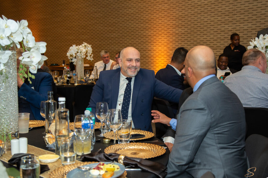 Smiling man in a blue suit sits at a formal dinner table with glassware and gold placemats on a dark tablecloth.