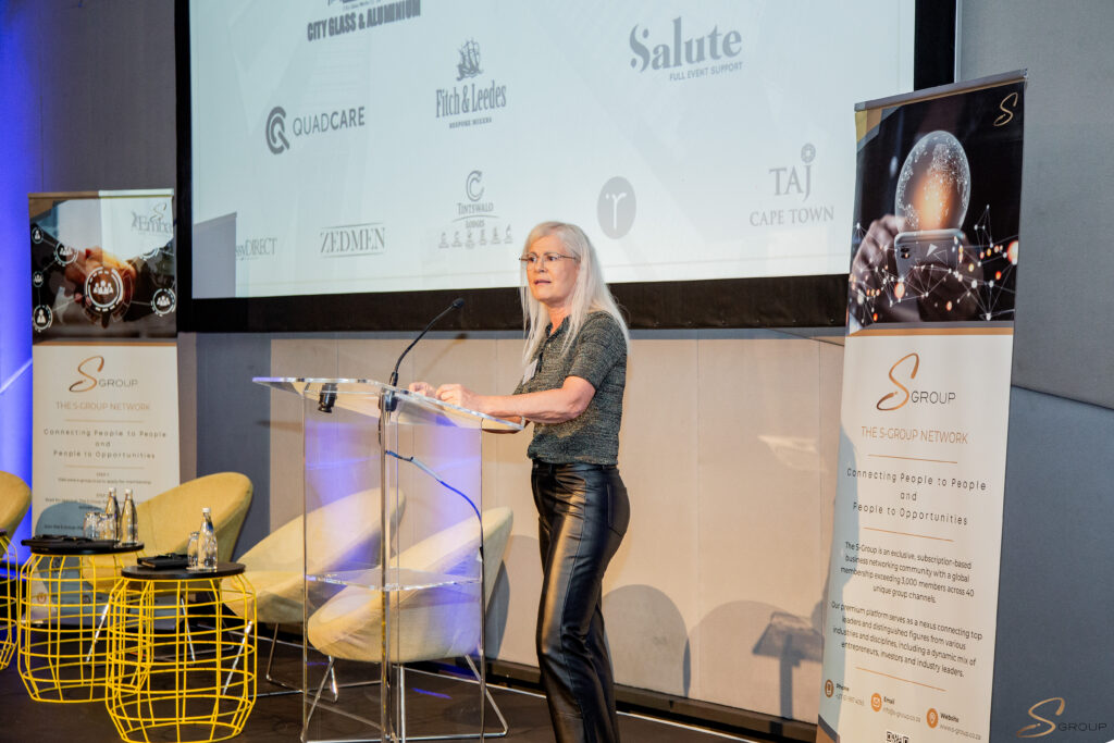 Woman with gray hair giving a talk at a clear podium on stage, banners and projection screen in the background.