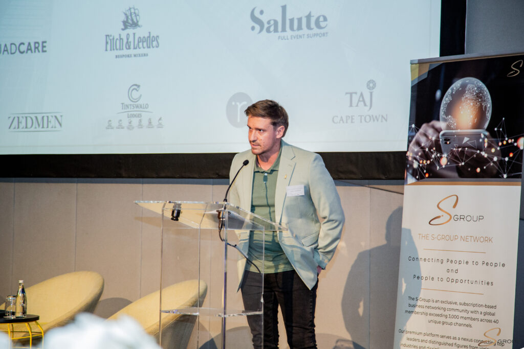 Man in a light blazer speaks from a clear acrylic podium at a conference, with sponsor logos on the backdrop behind him.