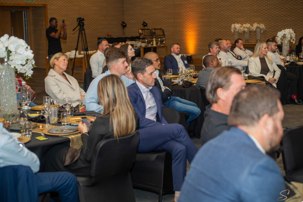 Professionals seated at round banquet tables in a conference room, listening to a presentation as a photographer records in the back area.