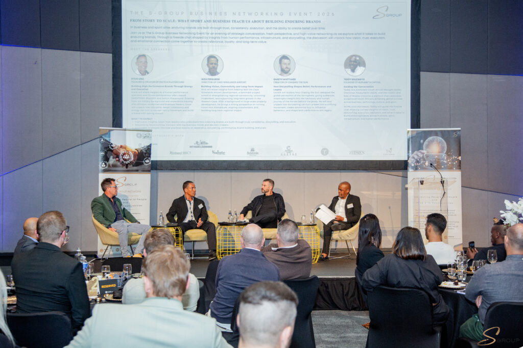 Panel discussion with four men seated on stage in a conference room, large screen banner behind them displaying event info.