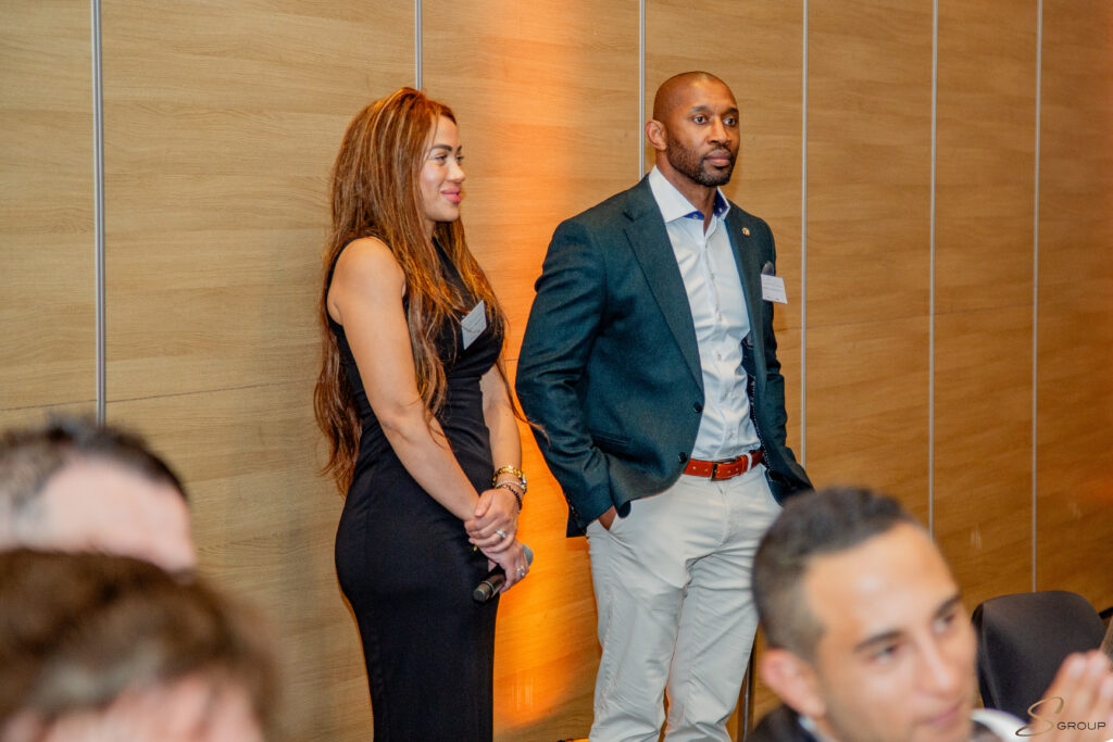 Two well-dressed individuals stand side by side at a formal event, a woman in a black dress on the left and a man in a blue blazer and white shirt on the right, with a wooden panel background and audience in the foreground.