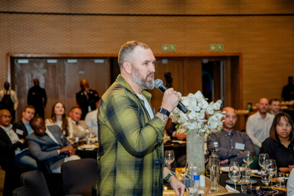 Man speaking into a microphone at a formal event, standing by a table with flowers and guests seated around him
