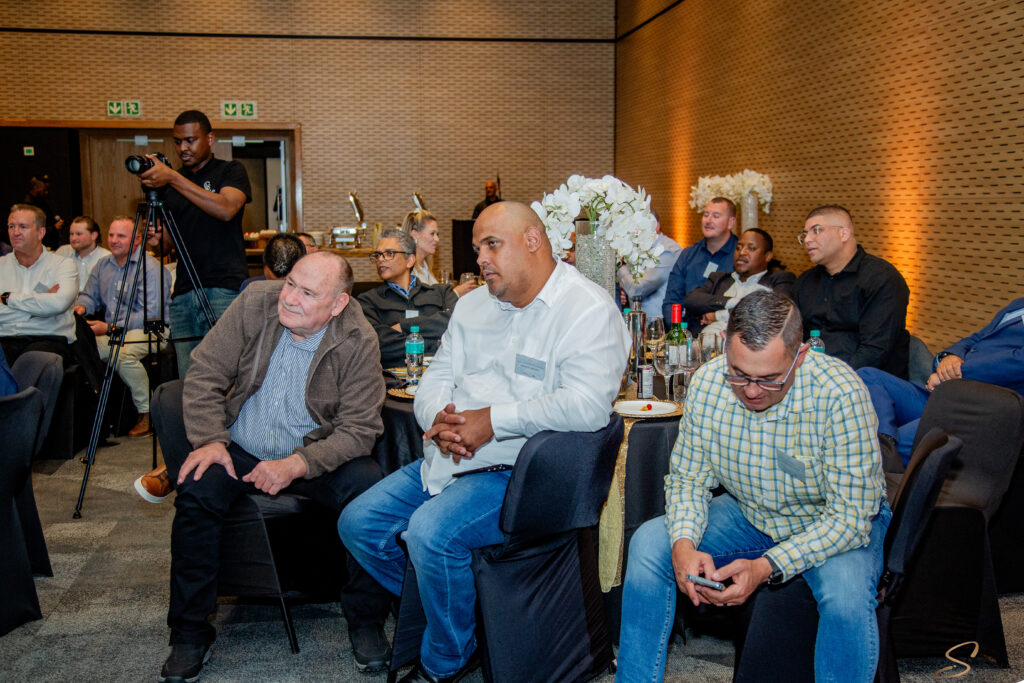 Audience at a banquet-style event with round tables and white floral centerpieces; a photographer on a tripod records the scene in the background.