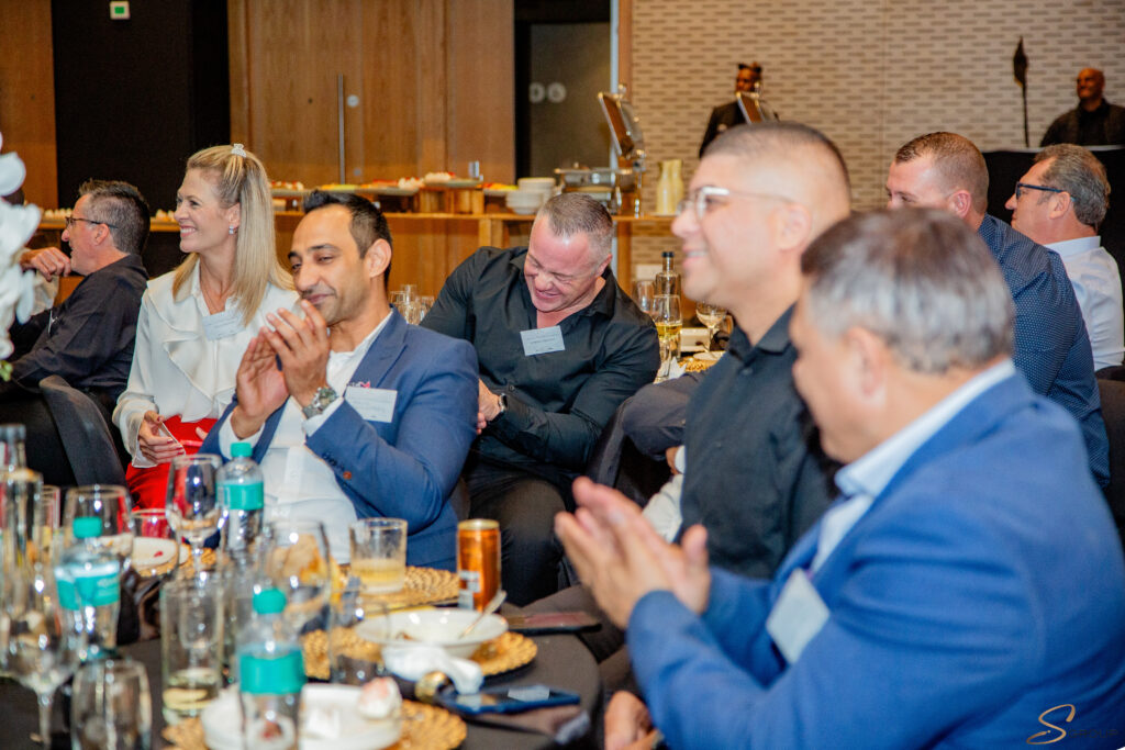 Group of adults in business attire seated at a banquet table, clapping and smiling during a formal event.