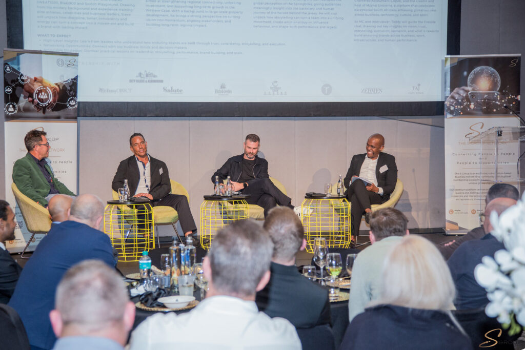 Four panelists sit on bright yellow wire-framed stools on a stage, engaged in discussion, with an audience in the foreground and a large projection screen and banners behind them.