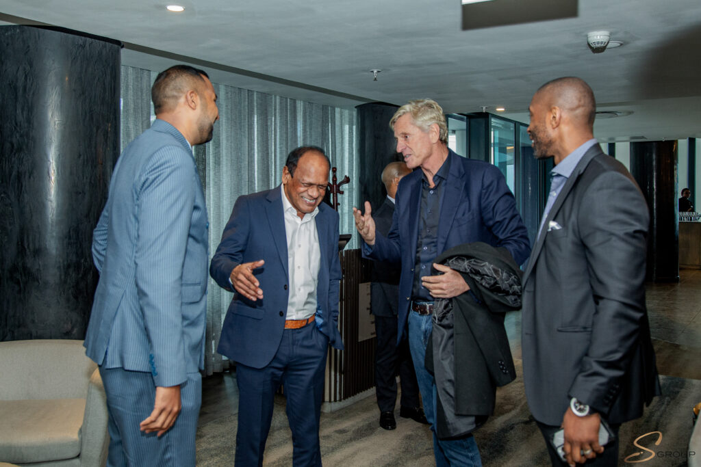 Four men in business attire conversing in a modern lobby with gray pillars and curtains in the background, smiling and gesturing as they talk.