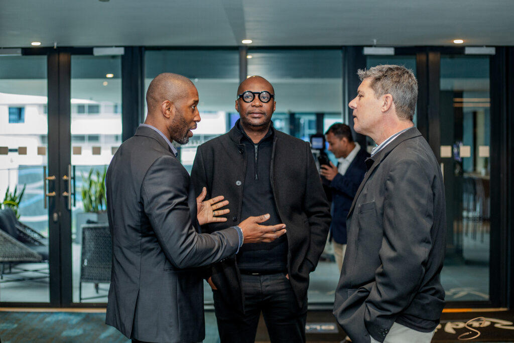 Three men in dark suits converse in a glass-walled office lobby with natural light.