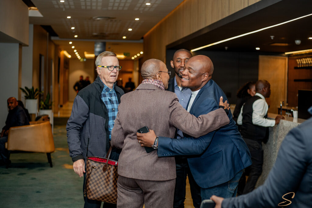 Group of people hugging warmly in a hotel lobby or reception area, smiling and greeting one another.