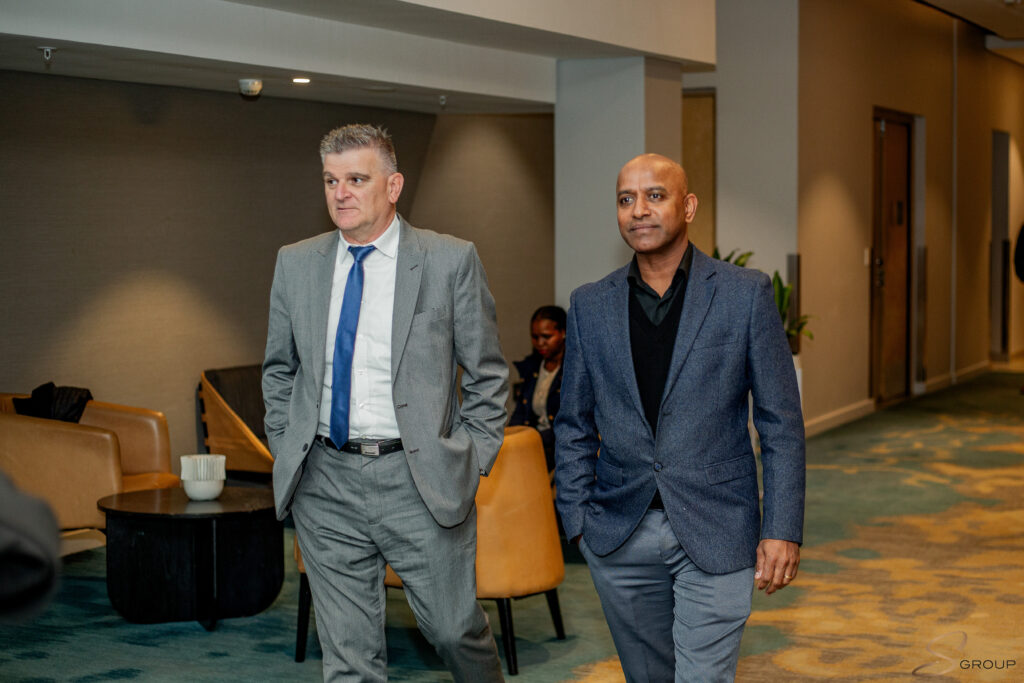 Two men in business suits walk together through a modern lobby area with seating and plants visible in the background.