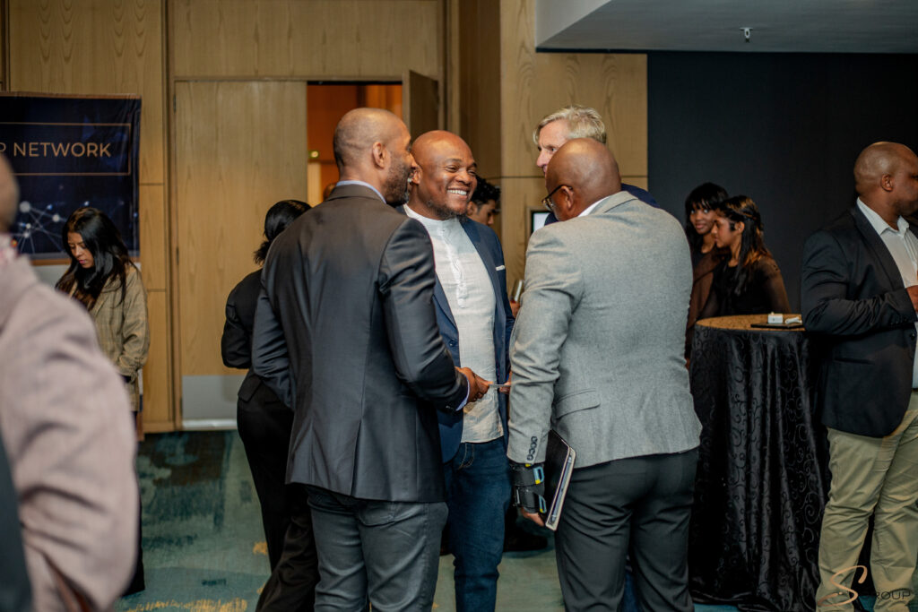 Professionals in suits converse and shake hands at a networking event in a wood-paneled lobby.
