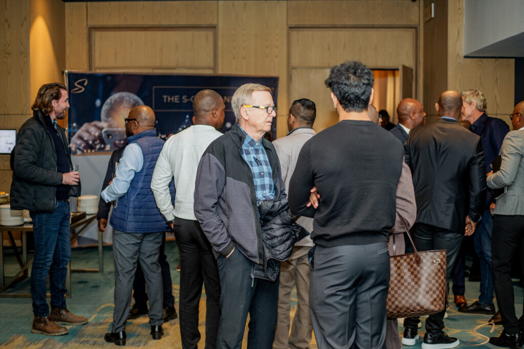 Group of men in business casual attire chatting at a crowded indoor event near a banner backdrop.