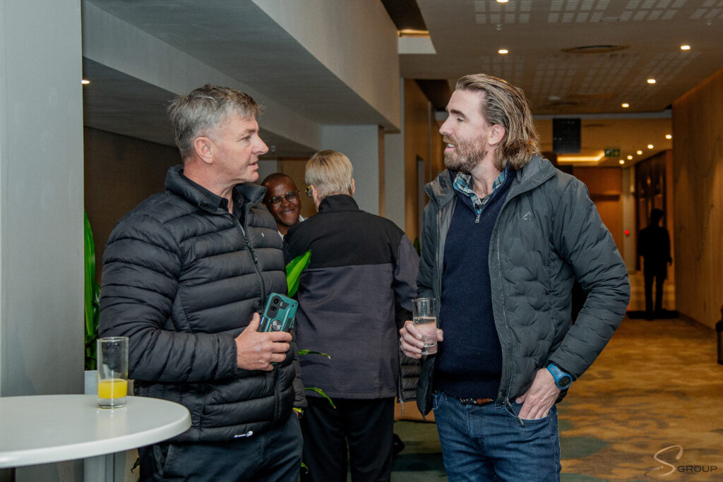 Two men in puffer jackets talk at a reception, one holding a smartphone and the other a glass of orange juice, with people chatting in the background.