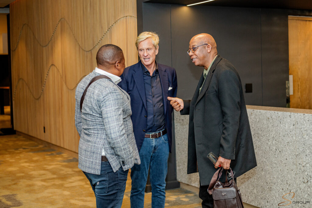 Three men in a hallway conversing; one in a navy suit, one in a gray plaid blazer, and a man in a dark overcoat holding a bag and glasses nearby.