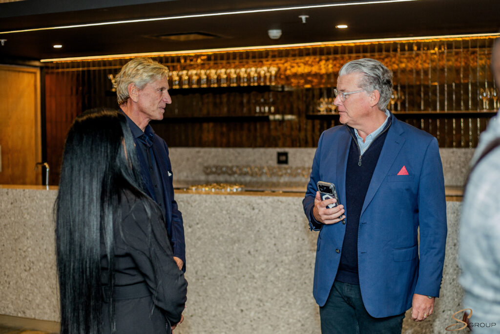 Two middle-aged men in a bar talking, one holding a smartphone and wearing a blue blazer while the other speaks nearby.