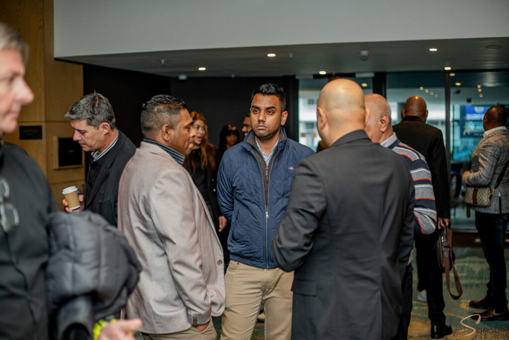 Group of professionals networking in a lobby; centered man in a blue quilted jacket looks toward the camera.Thinking/attentive expression.