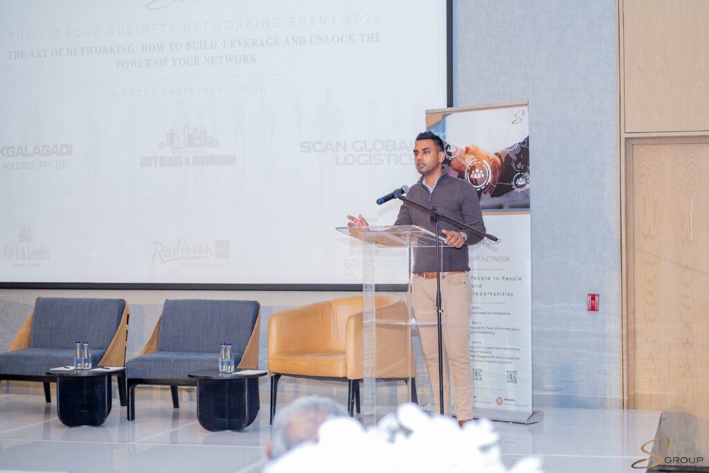 Speaker at a conference stands behind a clear lectern on stage, gesturing as he talks, with a banner and backdrop behind him.
