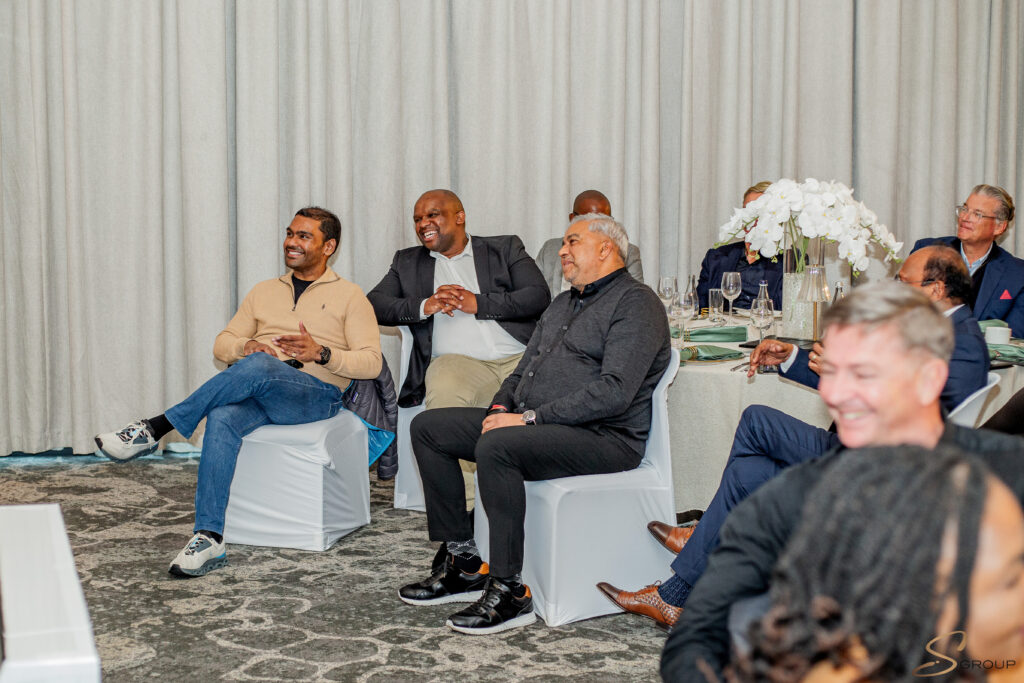 Six men sit in a row at a formal event, smiling and chatting, with a white floral centerpiece on the table behind them.