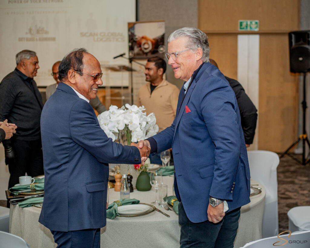 Two men in navy suits shake hands at a formal banquet with a flower centerpiece between them.