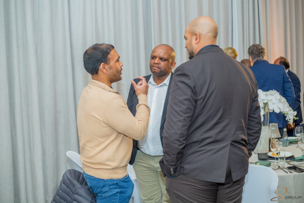 Three men in business casual conversing at a reception, with a table of drinks and flowers nearby against a light curtain backdrop.
