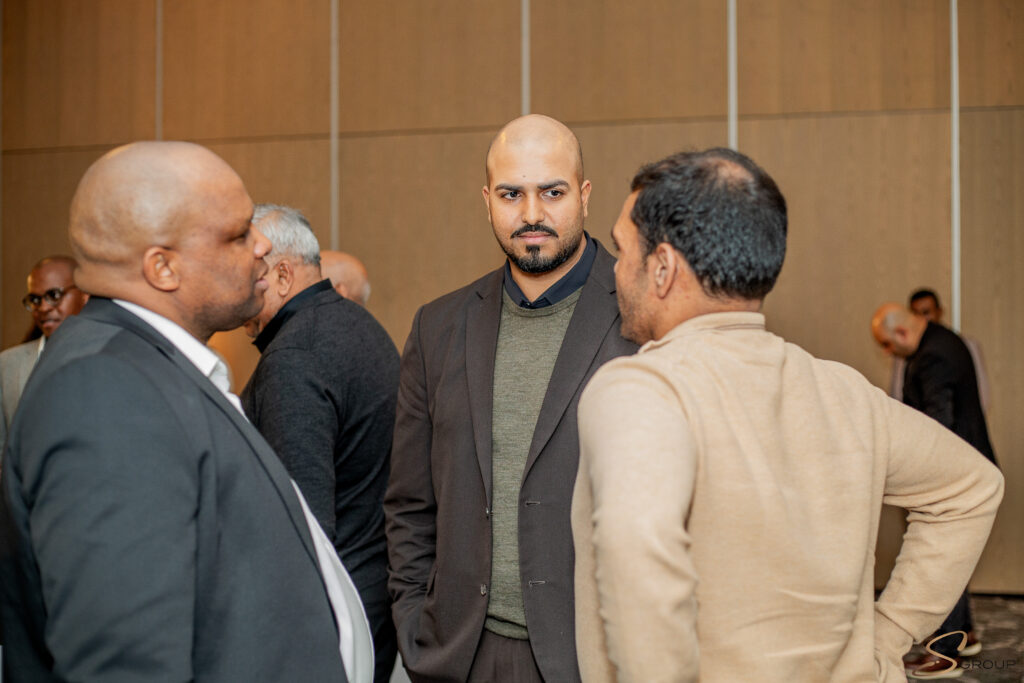 Man in a dark blazer and green sweater talks with others at a social/business gathering, wood-paneled wall behind them.