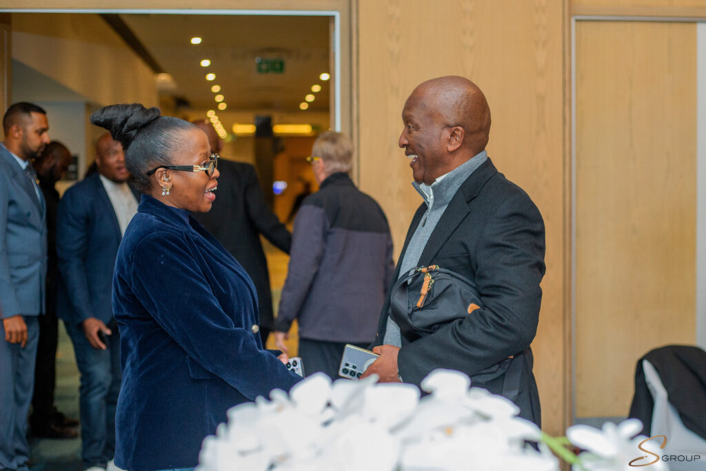 Two people shake hands and smile in a doorway, a woman in a blue velvet blazer and glasses facing a man in a dark jacket.