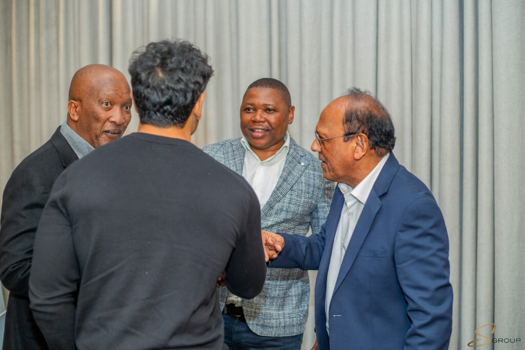 Group of four men in business attire chatting and shaking hands in a circles in front of a gray curtain backdrop, likely at a meeting or event.