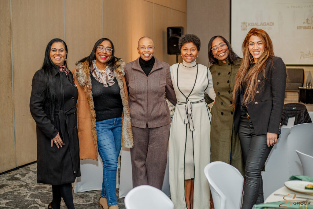 Six women in professional outfits smiling together at an indoor event/photo op.