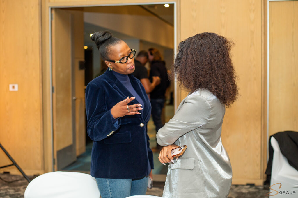 Two women face each other in a hallway, one in a dark blue velvet blazer with glasses, gesturing as she speaks while the other wears a silver-gray outfit and holds a phone nearby.