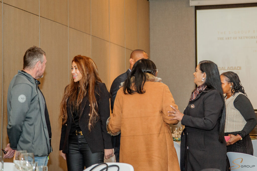 Group of professionals conversing at a networking event, standing along a beige paneled wall and smiling. (informative)