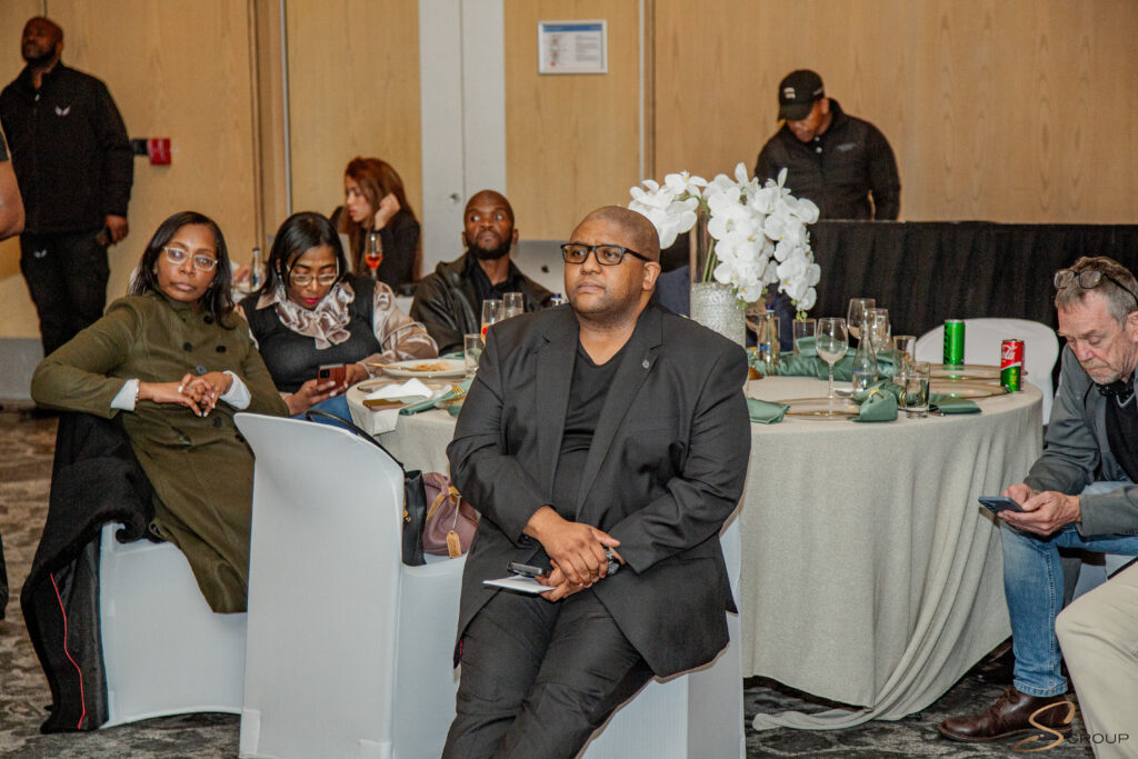 Man in a black suit with glasses sits at a round banquet table, hands clasped, with a white orchid centerpiece and other guests nearby.
