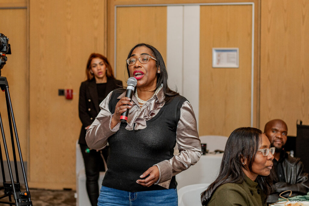 Woman speaking into a handheld microphone at a formal event, wearing a silver blouse and black vest, with other people and a camera in the background.