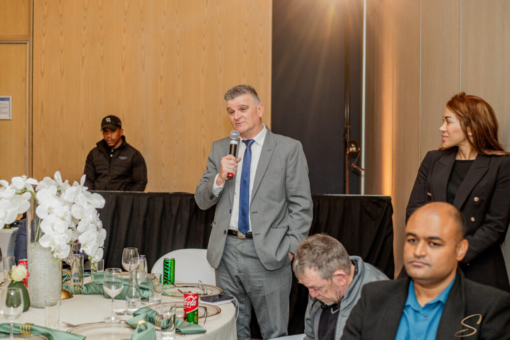 Man in a gray suit speaks into a handheld microphone at a banquet, standing beside a table with white orchids and place settings amid other guests in formal attire.