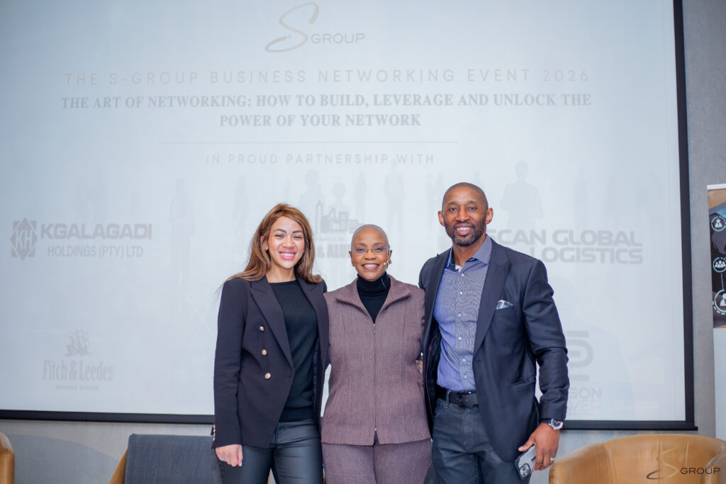 Three professionals pose together for a group photo at a business networking event, with a light backdrop showing logos and text in the background.