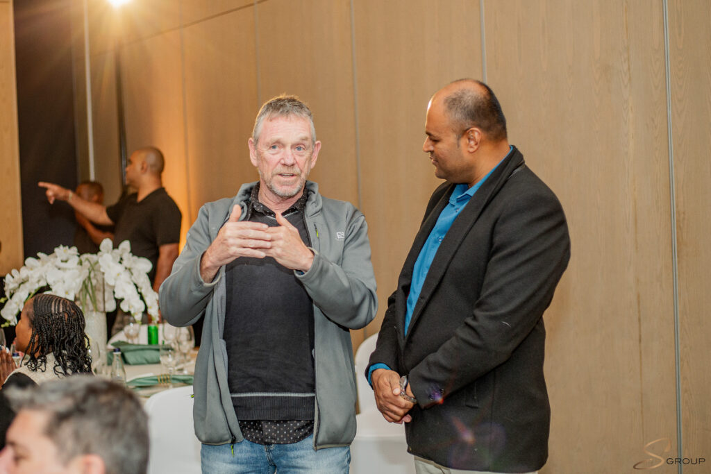 Two men stand and converse at a formal gathering, one gesturing with hands while the other listens nearby in a blazer and blue shirt, indoors beside a wooden wall.
