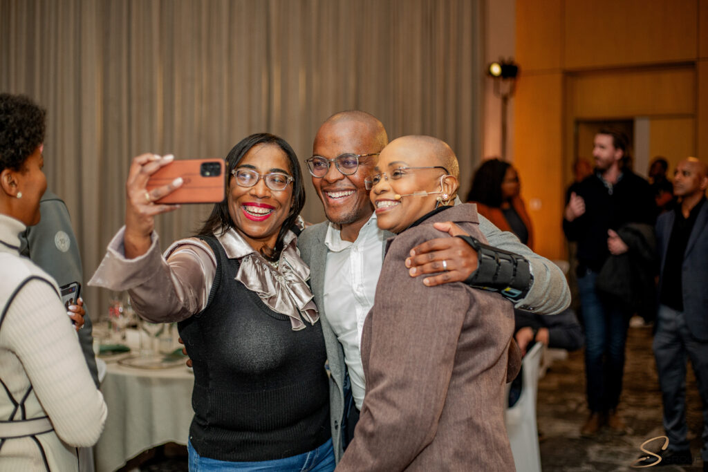 Three friends take a group selfie at a social event, with a smartphone held by the left person.